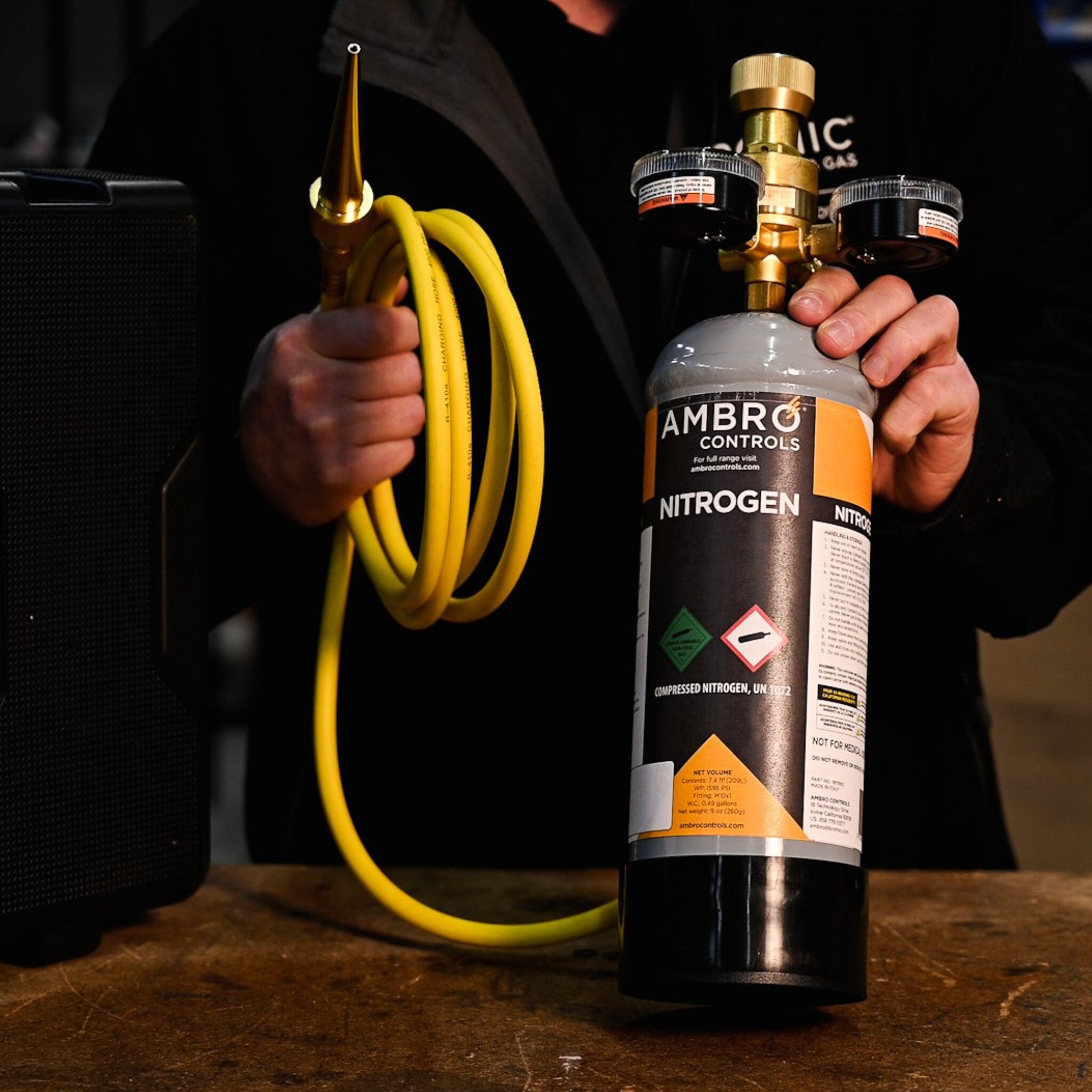 Technician holding a nitrogen cylinder in one hand and a coiled yellow hose with brass nozzle in the other, showcasing a portable purging setup.